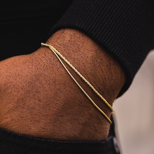 May include: Two gold chains, one a thin rope chain and the other a thicker, more textured rope chain, are worn on a person's wrist.