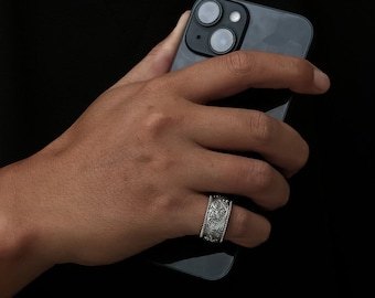 May include: A silver ring with an intricate floral design. The ring is worn on the finger of a person holding a black smartphone.