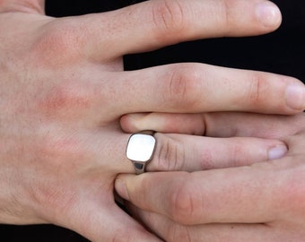 May include: A silver signet ring with a square face is worn on a finger. The ring has a polished, reflective surface. The background is a dark, solid color.