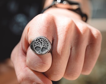 May include: A silver ring with a tree of life design. The ring is worn on the index finger of a hand.