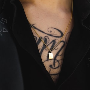 Könnte beinhalten: Eine goldene Kettenkette mit einem kleinen rechteckigen Anhänger. Die Kette wird von einer Person mit einem Tattoo auf der Brust getragen.