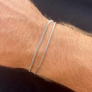 Pode incluir: Duas pulseiras de corrente prateadas, uma mais espessa que a outra, são usadas em um pulso. As pulseiras são feitas de pequenas elos interligados. O tom de pele é claro com pelos visíveis.