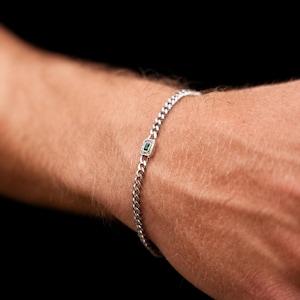 Pode incluir: Uma pulseira de prata com um design de corrente de elos. A pulseira apresenta uma pequena pedra preciosa verde em forma quadrada no centro. A pulseira é mostrada no braço de uma pessoa contra um fundo preto.