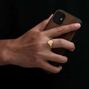 May include: A gold signet ring with a smooth oval top. The ring is worn on the index finger of a hand holding a wooden phone case.