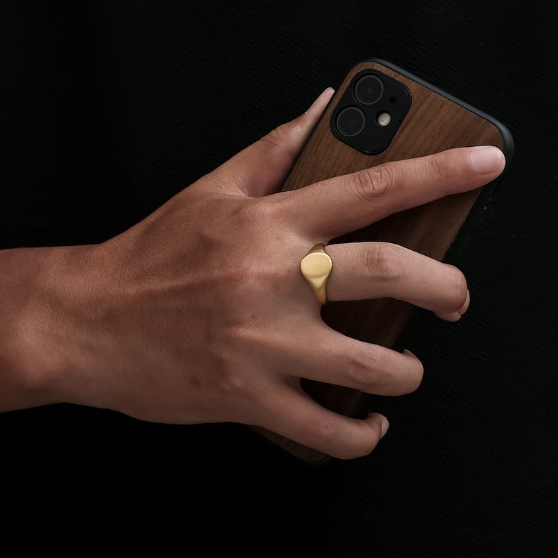 May include: A gold signet ring with a smooth oval top. The ring is worn on the index finger of a hand holding a wooden phone case.