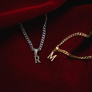 Puede incluir: Dos collares con iniciales sobre fondo rojo. Uno es plateado con la letra "R", el otro dorado con "M". Ambos tienen un diseño de cadena de eslabones.
