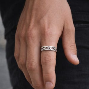 May include: A silver ring with a geometric diamond pattern. The ring is worn on a finger, showcasing its intricate design and metallic sheen. The ring's openwork design allows light to pass through, creating a visually appealing accessory.