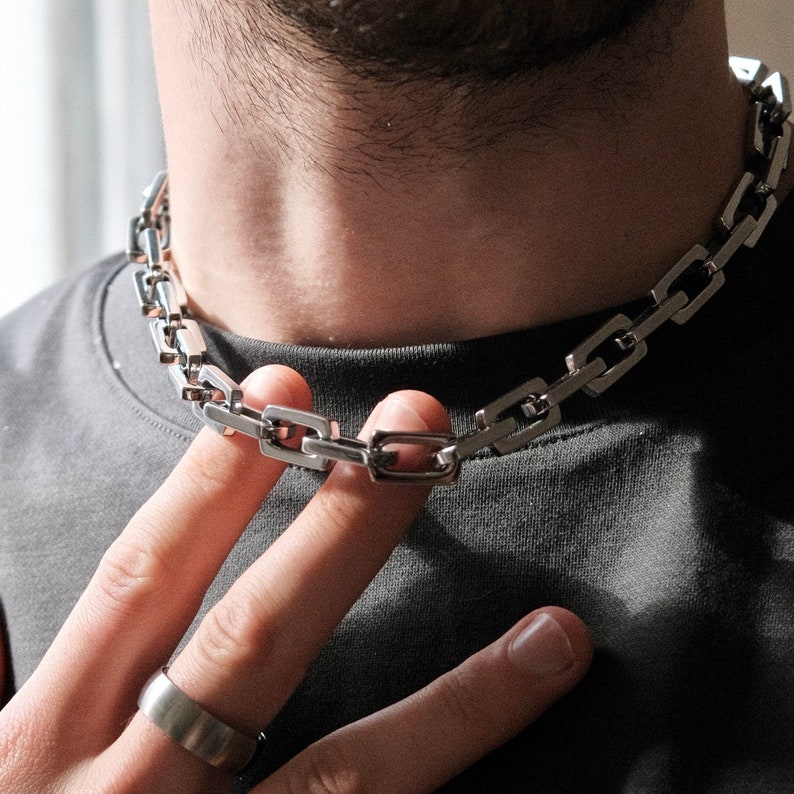 May include: A silver-colored chain necklace with rectangular links. The necklace is worn around the neck and is being held by a finger. A silver ring is also visible. The person is wearing a gray t-shirt.