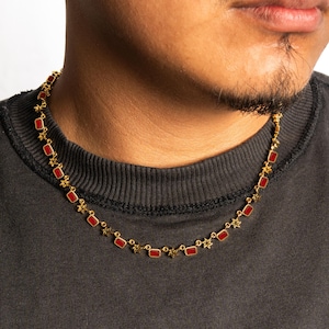 Puede incluir: Collar dorado con piedras rojas rectangulares. La cadena tiene detalles en forma de estrella entre cada piedra roja. El collar se lleva sobre una camiseta gris oscuro. Un accesorio elegante.