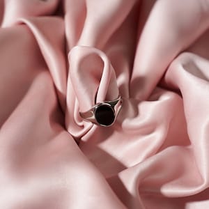 Op de afbeelding: Een zilveren ring met een zwarte, ovale steen. De ring is geplaatst op een achtergrond van zachte, roze satijnen stof. De band van de ring is glad en gepolijst. De algehele uitstraling is elegant en minimalistisch.