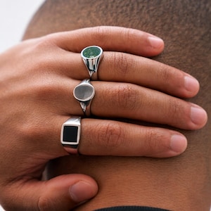 Puede incluir: Tres anillos de sello plateados con diferentes incrustaciones de piedra. El anillo superior tiene una piedra verde, el anillo central tiene una piedra gris y el anillo inferior tiene una incrustación cuadrada negra. Los anillos están en una mano.