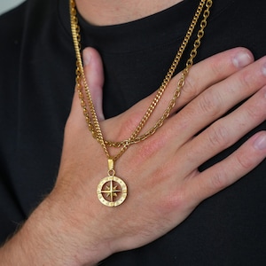 May include: Gold-coloured necklace with a compass pendant. The necklace features two chains, one with a curb link design and the other with a smaller link design. The compass pendant has a star design in the centre and is surrounded by a circular frame.