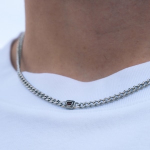 May include: Silver chain necklace with a rectangular, dark red gemstone accent. The necklace is a curb chain style, and is worn against a white t-shirt.
