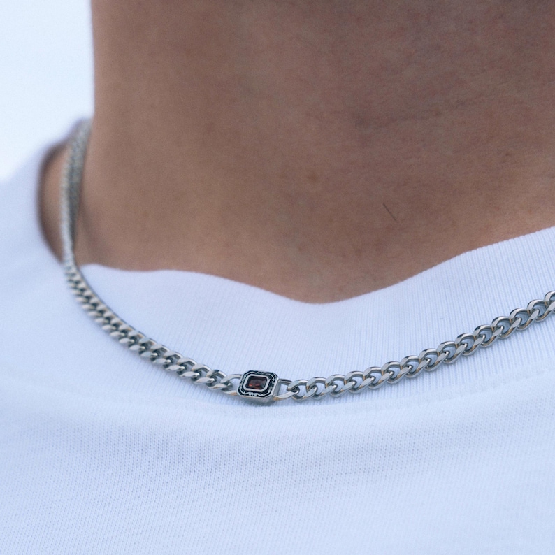 May include: Silver chain necklace with a rectangular, dark red gemstone accent. The necklace is a curb chain style, and is worn against a white t-shirt.
