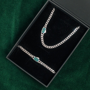 Può includere: Un set di collana e bracciale a catena argentata, ciascuno con un accento di pietra rettangolare color turchese. I gioielli sono presentati in una scatola nera su una superficie di velluto verde. Le maglie della catena sono ravvicinate.