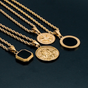 Op de afbeelding: Vier gouden kettingen met verschillende hangers. De hangers omvatten een zwart vierkant, een ronde hanger met een zwart midden, een ronde hanger met een kompasontwerp en een ronde hanger met een religieuze figuur.