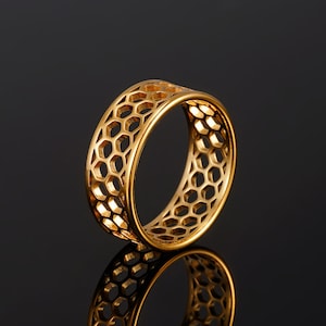 May include: A gold ring with a honeycomb pattern. The ring is a band style, with a series of interconnected hexagonal shapes. The ring is shown on a reflective surface, creating a mirrored effect. The ring is a piece of jewellery.
