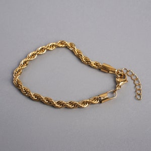 May include: Gold-colored rope chain bracelet. The bracelet features a textured, twisted rope design. It has a lobster clasp closure and an adjustable chain. The bracelet is displayed on a gray background.