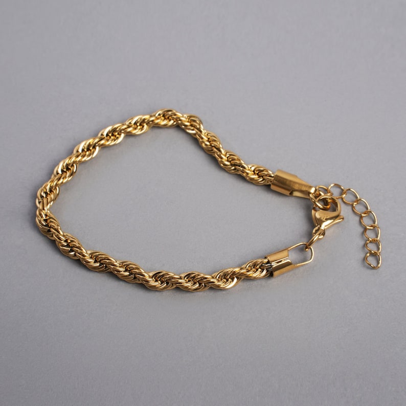 May include: Gold-colored rope chain bracelet. The bracelet features a textured, twisted rope design. It has a lobster clasp closure and an adjustable chain. The bracelet is displayed on a gray background.
