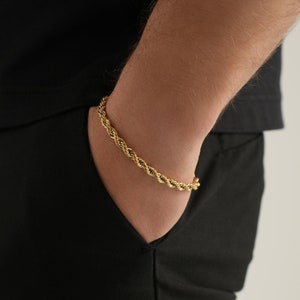 May include: A gold-colored rope chain bracelet. The bracelet is made of twisted metal links, creating a rope-like texture. The bracelet is worn on a wrist, with a black shirt and black pants in the background.