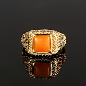 Puede incluir: Un anillo de oro con una piedra preciosa naranja engastada en un bisel cuadrado. El anillo tiene un diseño intrincado con motivos florales.