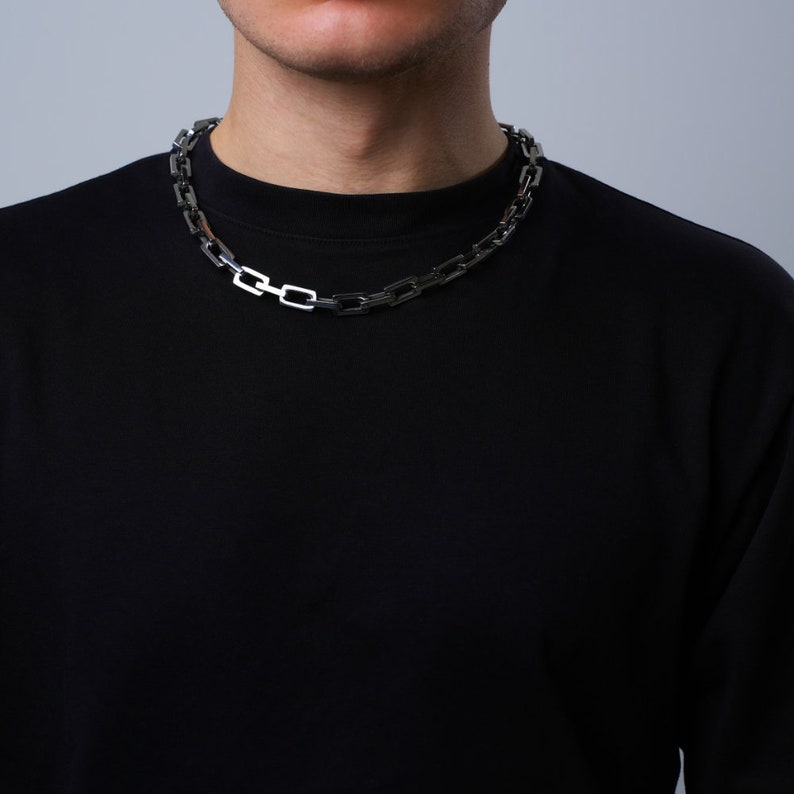 May include: A silver-colored chain necklace with a rectangular link design. The necklace is worn over a black crew neck shirt. The background is a solid light gray.