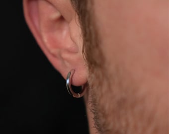 May include: A silver hoop earring is worn in the ear. The earring is a simple, circular design with a polished, reflective surface. The earring is a classic accessory.
