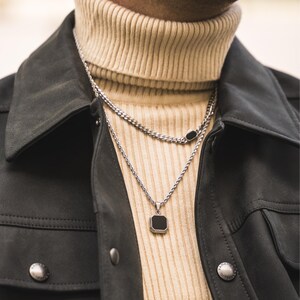 May include: A silver necklace with a square black pendant and a second chain necklace with a black oval accent. The necklaces are layered over a beige turtleneck sweater and a dark gray jacket.