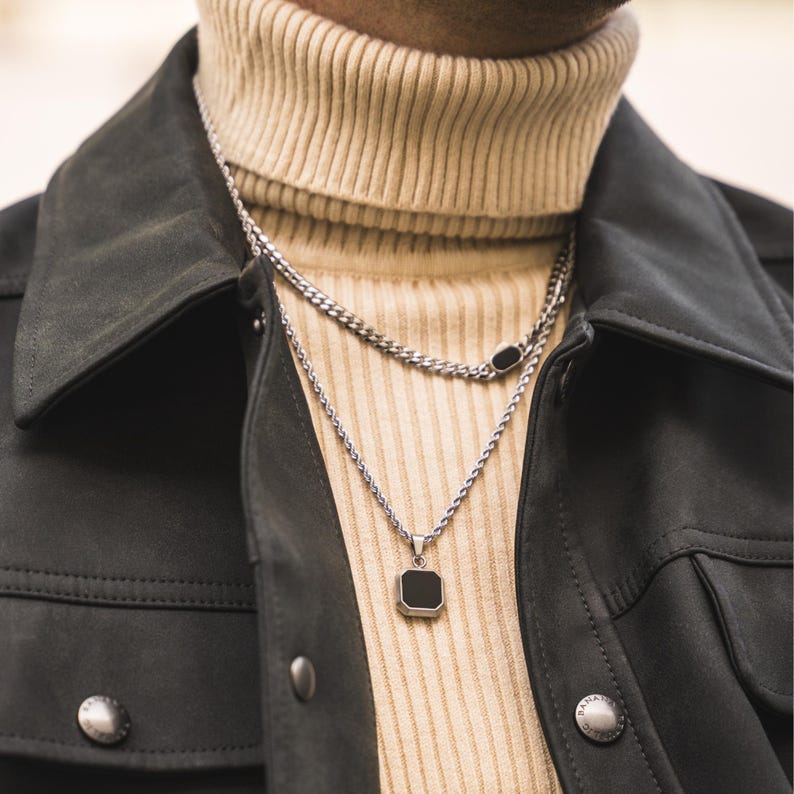 May include: A silver necklace with a square black pendant and a second chain necklace with a black oval accent. The necklaces are layered over a beige turtleneck sweater and a dark gray jacket.
