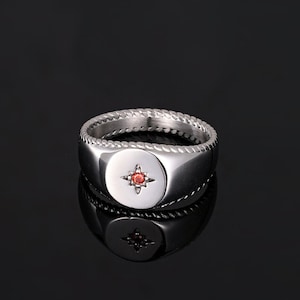 May include: A silver signet ring with a red starburst design set in a circular face. The ring has a braided rope-like texture around the band. The ring is displayed on a reflective black surface.
