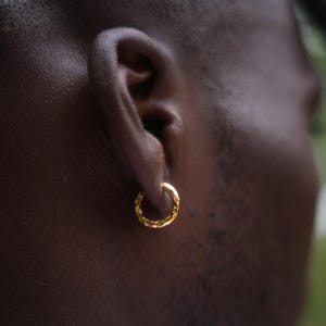 May include: Close-up of a gold hoop earring with a textured surface. The earring is circular and appears to be made of metal. The earring is worn on the ear of a person with dark skin.