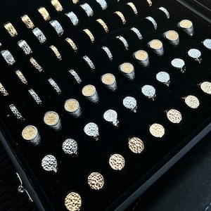May include: A selection of rings presented in a black velvet jewellery box. The rings have diverse designs, including textured bands and circular faces with gold or silver finishes. The display highlights a range of styles and sizes.