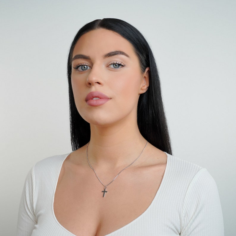 May include: A woman with long black hair wearing a white shirt and a silver necklace with a small cross pendant.