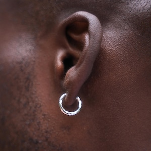 May include: Close-up of a silver hoop earring with a textured surface. The earring is circular and hangs from an ear. The skin tone is dark.