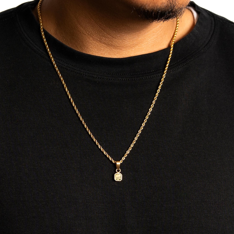 May include: Gold-colored rope chain necklace with a small, square pendant. The pendant has a light-colored stone in the center. The necklace is worn against a black shirt.