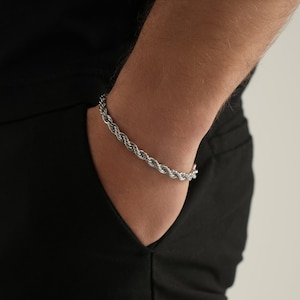 May include: A silver-colored rope chain bracelet. The bracelet is made of tightly woven metal strands, creating a textured, braided appearance. The bracelet is worn on a wrist, against a black background.