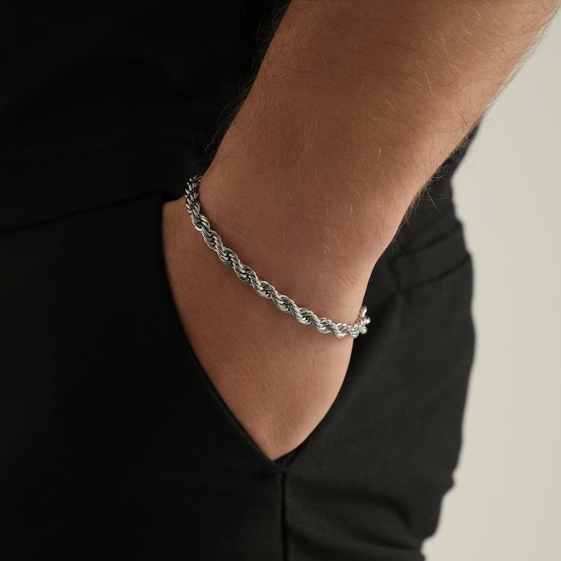 May include: A silver-colored rope chain bracelet. The bracelet is made of tightly woven metal strands, creating a textured, braided appearance. The bracelet is worn on a wrist, against a black background.