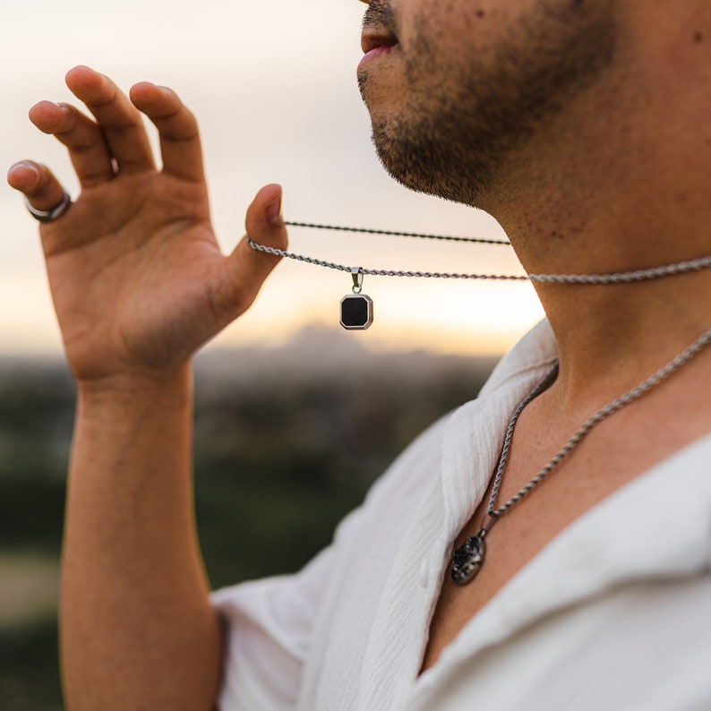 May include: A silver necklace with a square black pendant. The pendant is held up by a hand. The image shows a person wearing a white shirt and other silver necklaces. The background is blurred with a sunset.