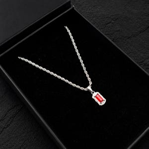 May include: A silver necklace with a rectangular red pendant, framed by small clear stones, sits in a black jewellery box. The chain has a twisted rope design. The box is open, displaying the necklace against a dark backdrop.