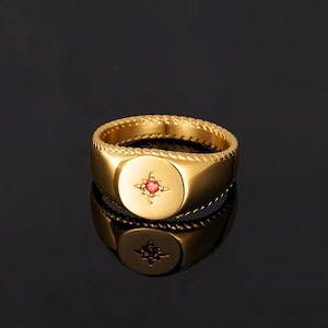 May include: A gold signet ring featuring a red gemstone in a starburst design. The ring has a braided band and a polished finish. It is shown on a reflective black surface. The ring's design is classic.