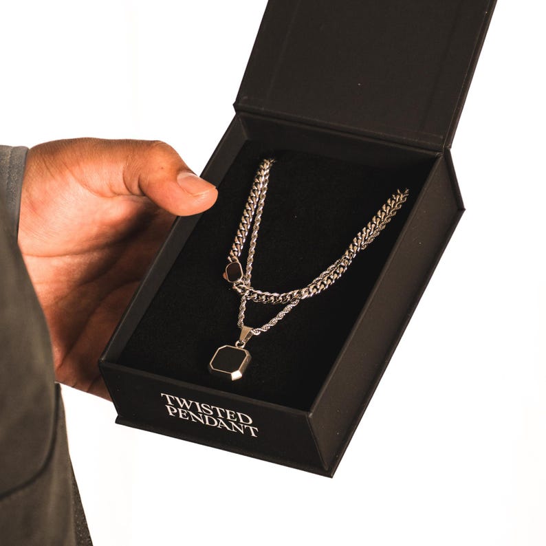 May include: A silver necklace with a black square pendant is displayed in a black box. The box is open, revealing the jewelry against a black velvet interior. The box has the words "TWISTED PENDANT" printed on the front.