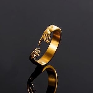 May include: A gold-toned open ring featuring two sculpted hands reaching toward each other at the opening. The band has a smooth, polished finish on the interior and a detailed, textured design of fingers and palms on the exterior ends.