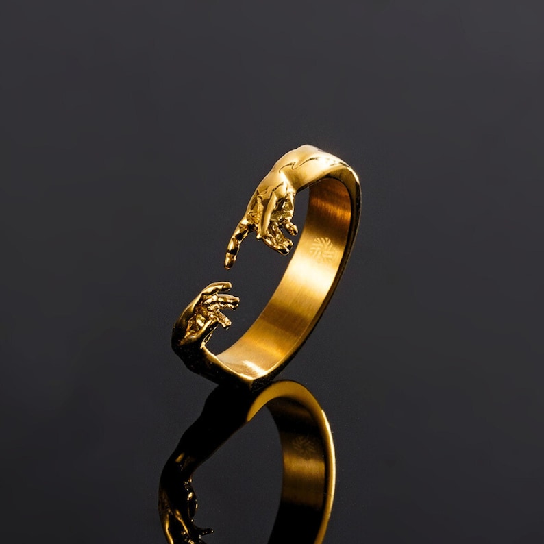 May include: A gold-toned open ring featuring two sculpted hands reaching toward each other at the opening. The band has a smooth, polished finish on the interior and a detailed, textured design of fingers and palms on the exterior ends.
