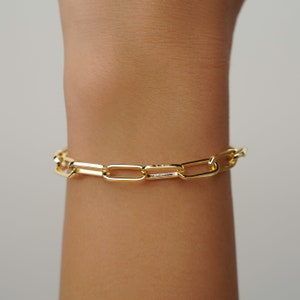 May include: A gold-colored link chain bracelet. The bracelet features a series of rectangular links, creating a modern and elegant design. The bracelet is worn on a wrist, showcasing its polished finish and simple, yet stylish aesthetic.
