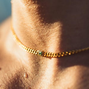May include: A gold choker necklace with a small square emerald-colored stone in the center. The necklace has a textured chain design and is worn around the neck. The image is a close-up shot.