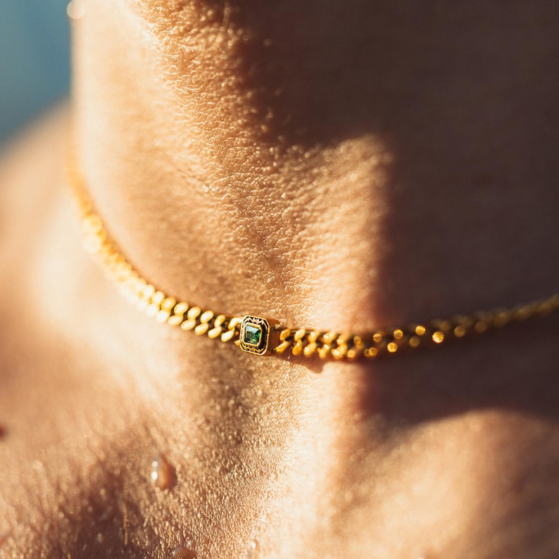 May include: A gold choker necklace with a small square emerald-colored stone in the center. The necklace has a textured chain design and is worn around the neck. The image is a close-up shot.