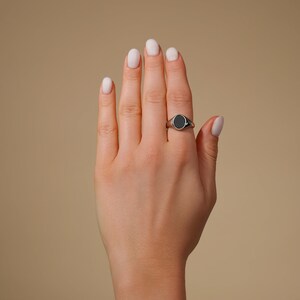 Op de afbeelding: Een zilveren zegelring met een zwarte ovale steen wordt gedragen op een hand met verzorgde nagels. De ring bevindt zich op de ringvinger en toont de gepolijste metalen band en donkere steen. De achtergrond is effen en neutraal.