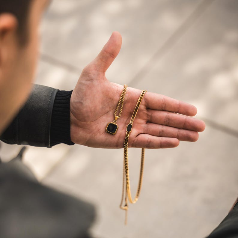 May include: Gold-colored necklaces with black accents. One necklace features a square pendant with a black stone, while the other has a rectangular black stone. The chains are displayed on a hand.