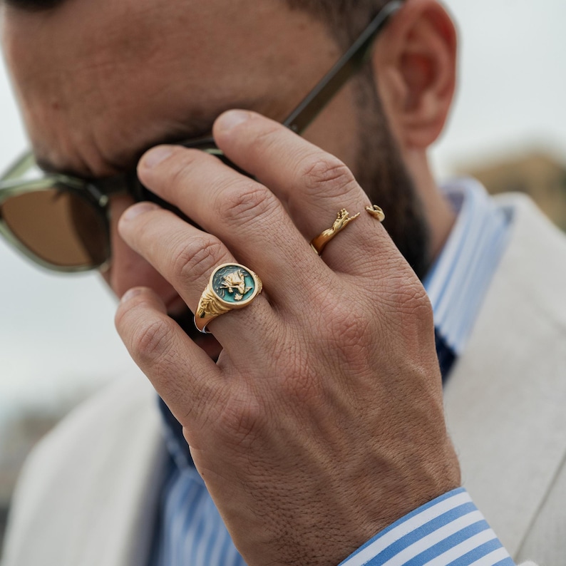 May include: A gold signet ring featuring a circular face with a teal inlay and a raised gold emblem, worn alongside a second gold ring designed as a pair of reaching hands. Both rings are crafted from polished gold metal with intricate detailing.