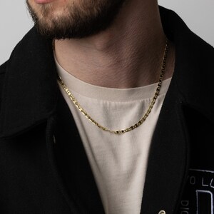 May include: A gold chain necklace with a mariner link design. The chain is worn by a person wearing a black jacket and a beige t-shirt.